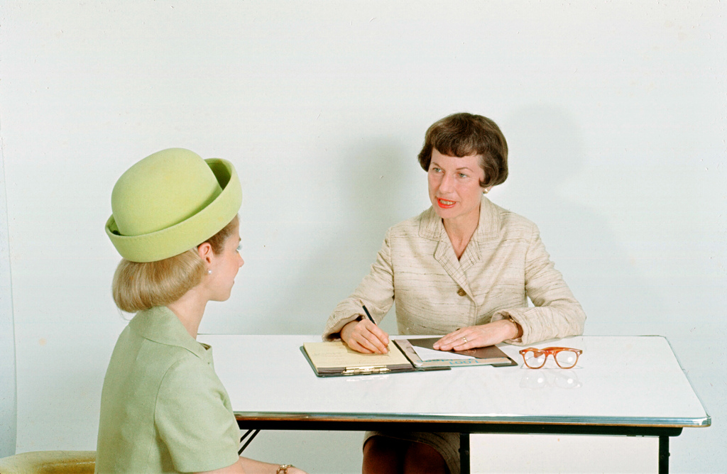 A young woman sits accross from an older woman who has a notebook. Both are wearing old fashioned dress. It looks like a job interview.